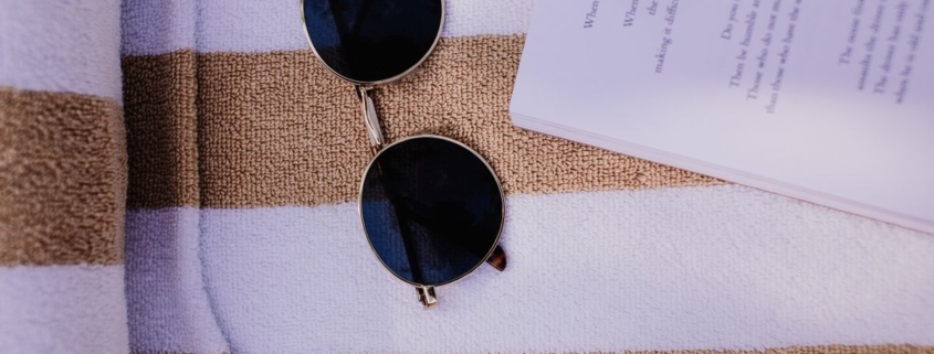 Open boek en zonnebril op een strandhanddoek, vakantiethrillers van Suzanne Vermeer